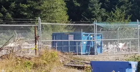 A blue trash can located behind a chain link fence, creating a contrast with the surrounding environment.